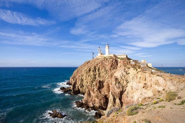 Un faro bianco e edifici si ergono su una scogliera aspra e rocciosa che si affaccia sul mare blu profondo sotto un cielo parzialmente nuvoloso.