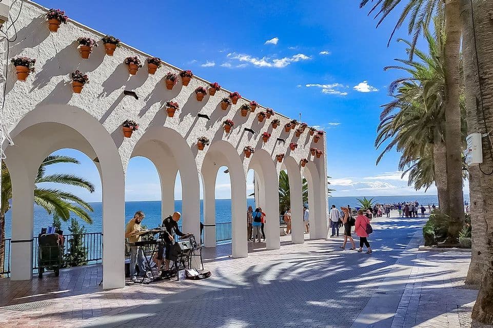 Due musicisti di strada suonano le tastiere sotto un colonnato bianco e ad arco, su una passeggiata marittima assolata con palme.