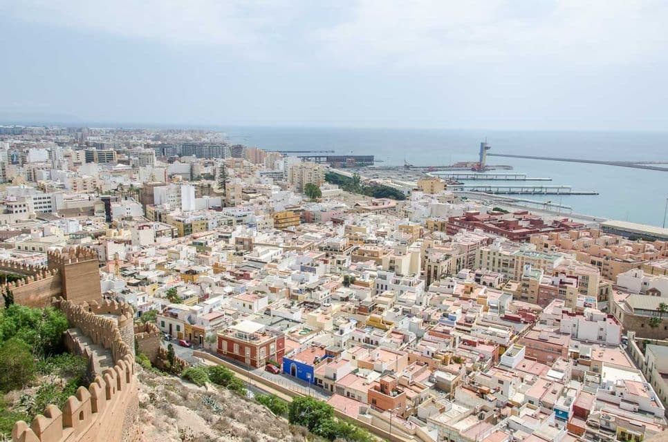 Una vista aerea di una città costiera densamente popolata, con un'antica cinta muraria fortificata in primo piano e un grande porto sullo sfondo.