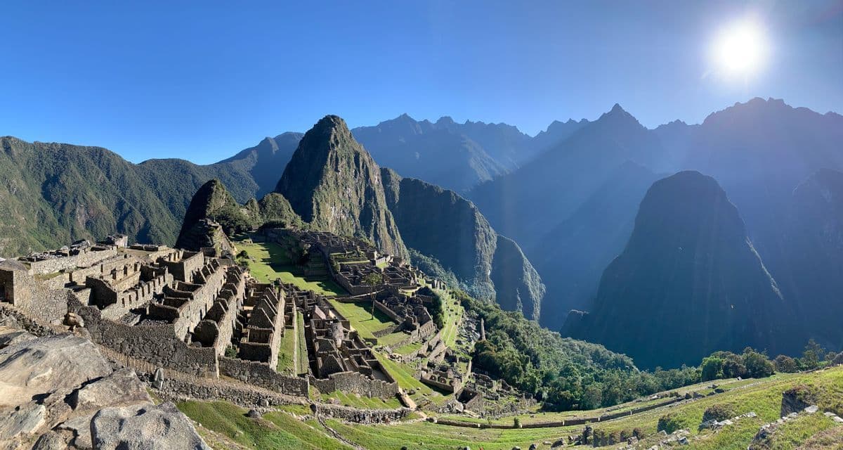 Panoramablick auf antike Steinruinen an einem terrassierten Berghang, umgeben von steilen Gipfeln unter klarem Himmel.