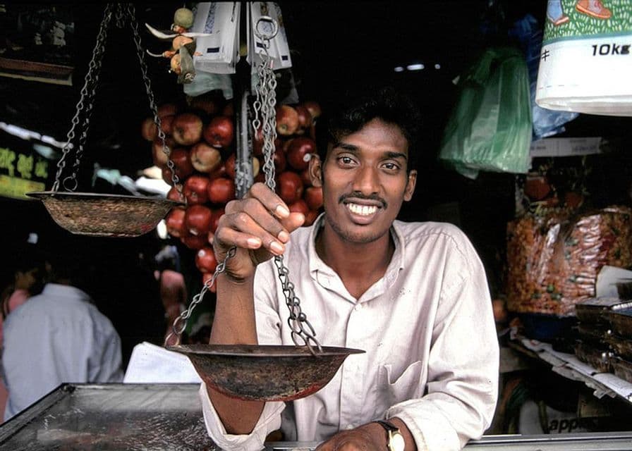 Un vendedor sonríe mientras sostiene una balanza tradicional en un puesto de mercado con manzanas colgando de fondo.