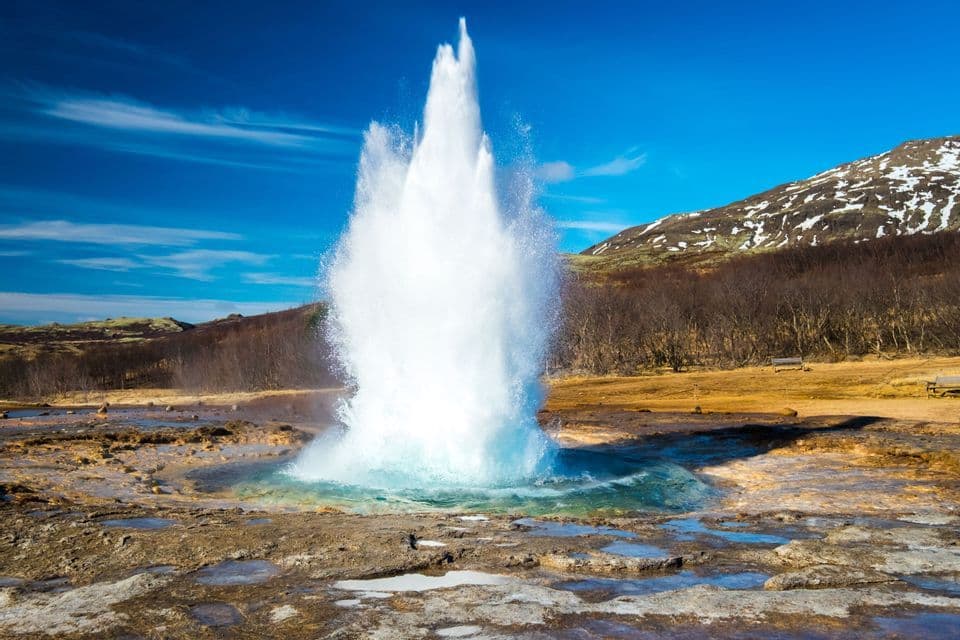 Un geyser jaillit d'une piscine turquoise, projetant un puissant panache d'eau dans les airs sur fond de collines saupoudrées de neige.