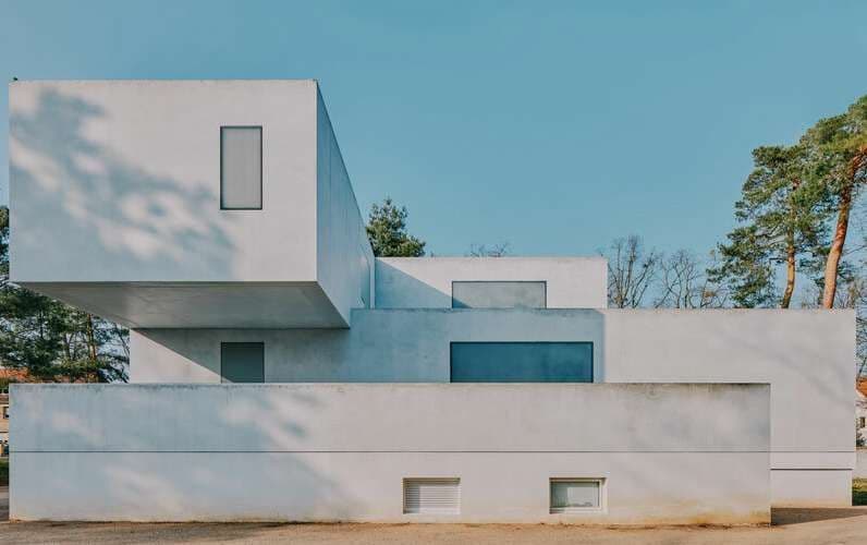 Un edificio bianco dal design minimalista e geometrico, con una sezione a sbalzo contro un cielo azzurro limpido.