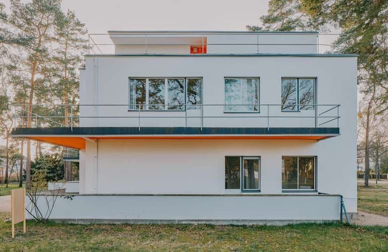Una moderna casa bianca con tetto piatto e un balcone prominente con la parte inferiore arancione, su uno sfondo di alberi alti.