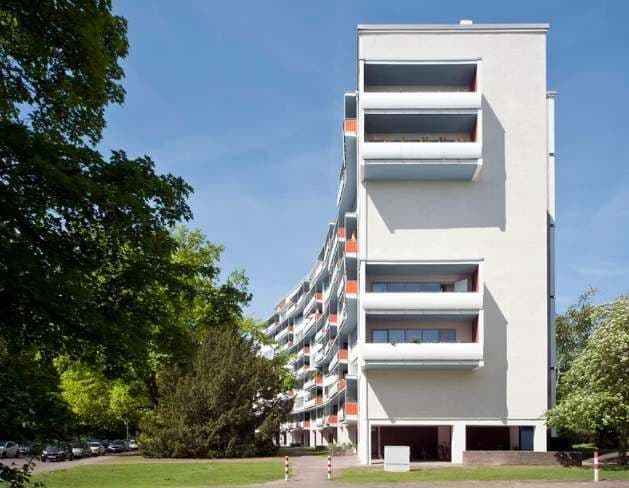 Un edificio residenziale curvo e bianco con balconi sorge accanto a un parco con alberi verdi e lussureggianti sotto un cielo azzurro e limpido.