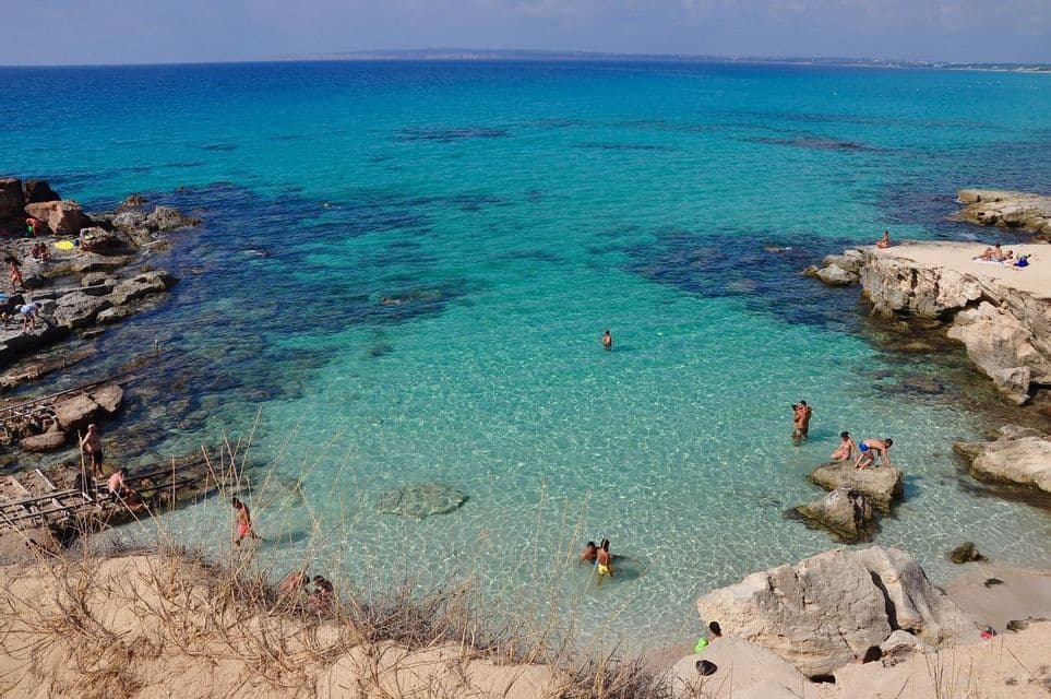 Gente nadando y tomando el sol en una cala rocosa con agua clara y turquesa bajo un cielo soleado.