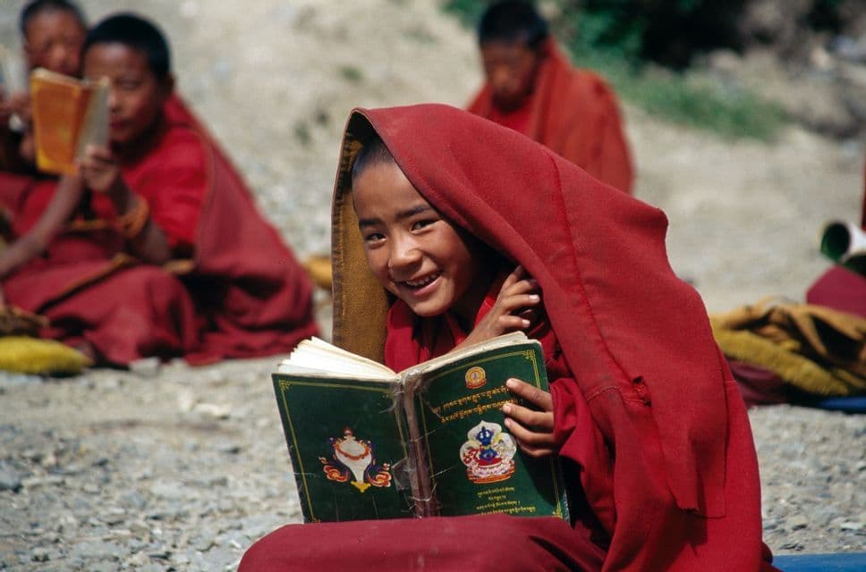 Un joven monje con una túnica roja con capucha sonríe mientras sostiene un libro abierto, sentado al aire libre con otros monjes al fondo.