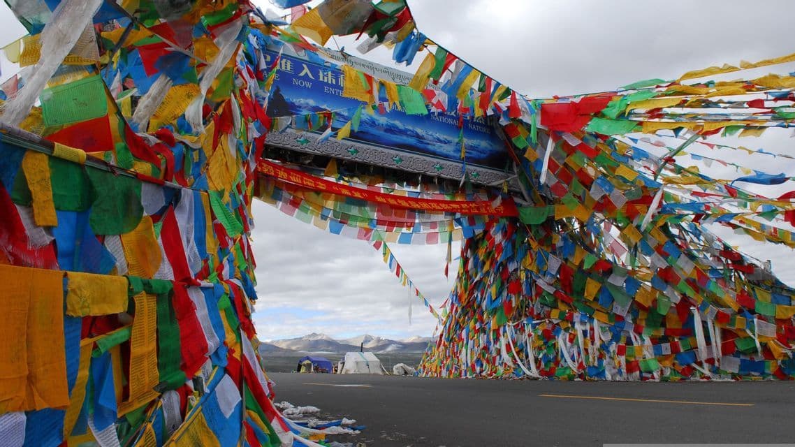 Un arco cubierto de coloridas banderas de oración cruza una carretera, con montañas distantes visibles bajo un cielo nublado.