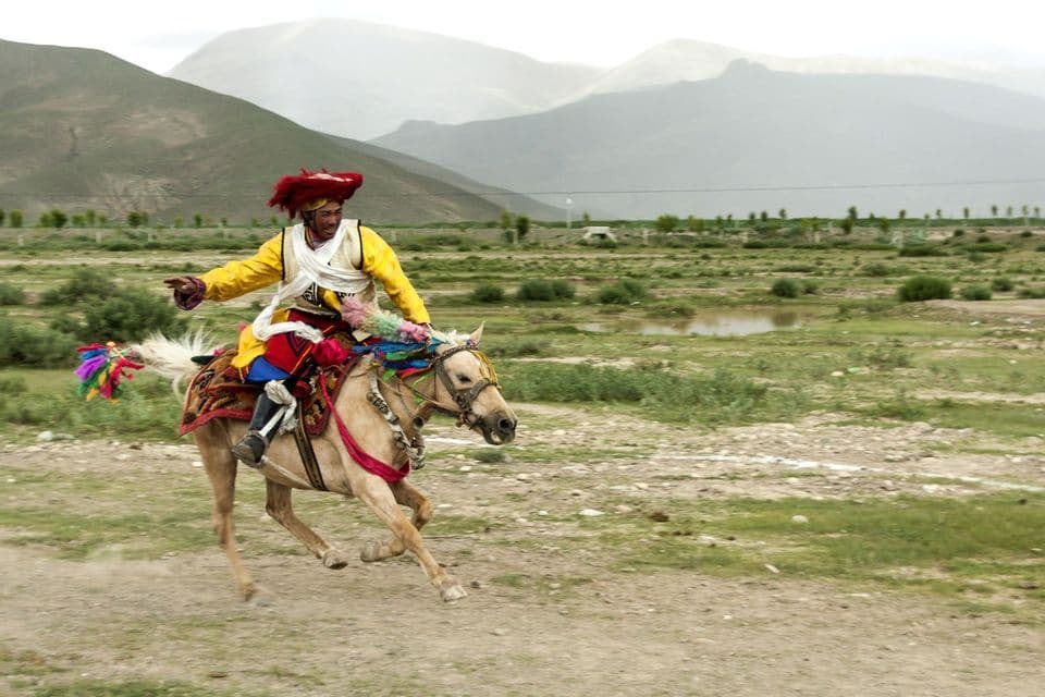 Un hombre con un traje tradicional amarillo y rojo cabalga un caballo decorado al galope por un campo de hierba con montañas brumosas detrás.