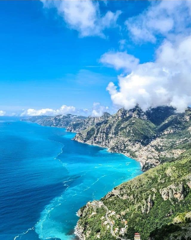 Eine Vogelperspektive auf eine bergige, grüne Küstenlinie, die an ein lebhaftes türkises und blaues Meer unter einem teilweise bewölkten Himmel grenzt.