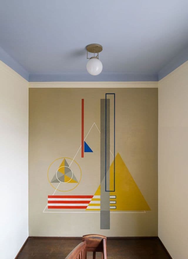 Un murale geometrico astratto con colori primari su una parete del corridoio, situato sotto un soffitto blu e una lampada a sospensione sferica.