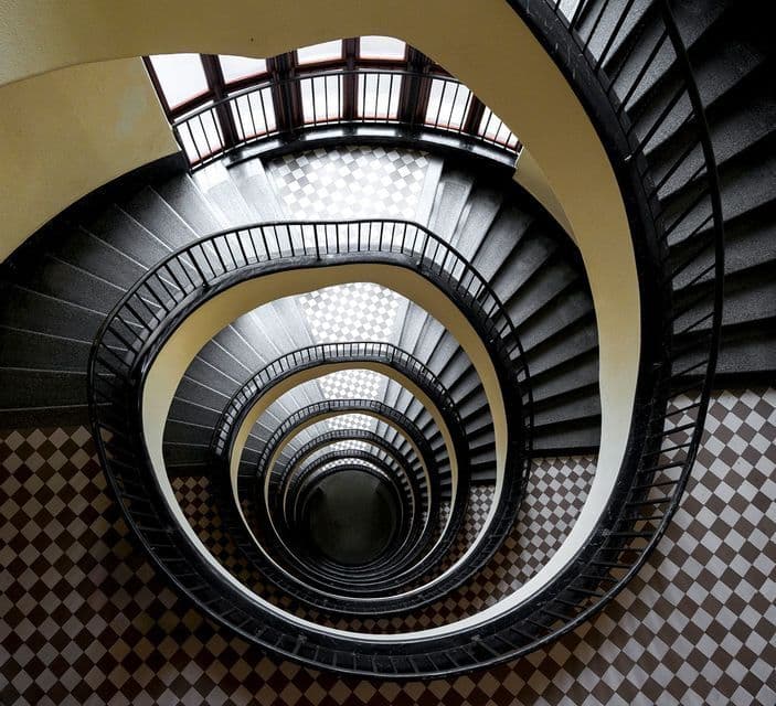 Vista dall'alto di una scala a chiocciola con ringhiera nera, che crea un motivo a vortice su un pavimento a scacchiera.