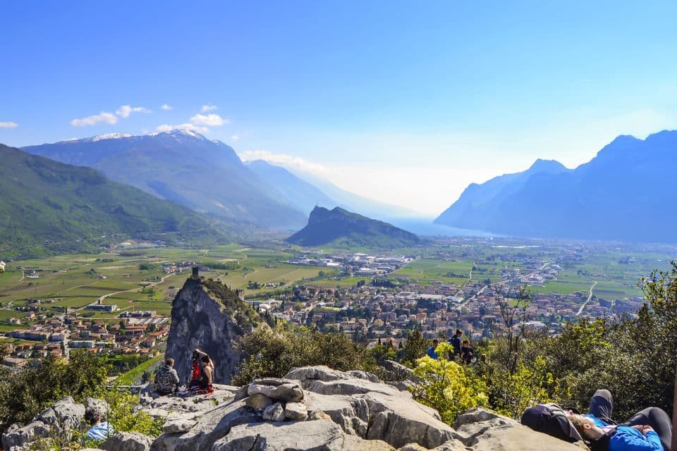 Un gruppo WeRoad si riposa su uno sperone roccioso, godendosi la vista panoramica di una valle, un lago e montagne con cime innevate.