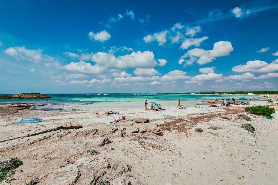 Bagnanti su una spiaggia sabbiosa con tratti rocciosi, accanto ad acque turchesi sotto un cielo azzurro con nuvole.