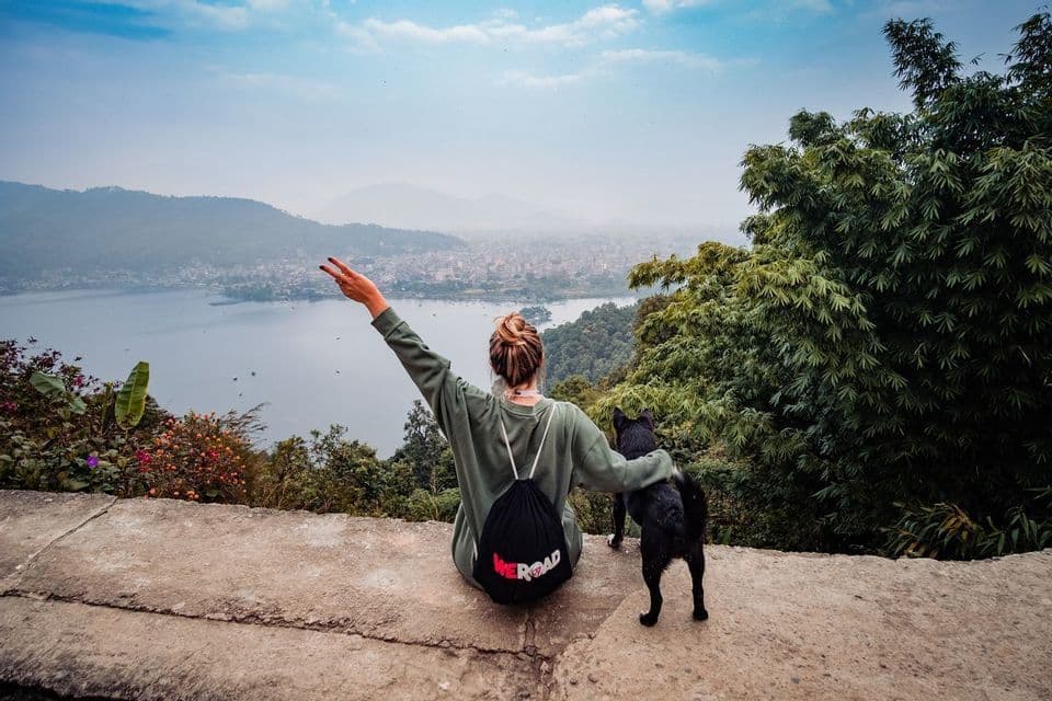 A person with a WeRoad backpack sits with a dog on a ledge, making a peace sign over a lake and a distant city.