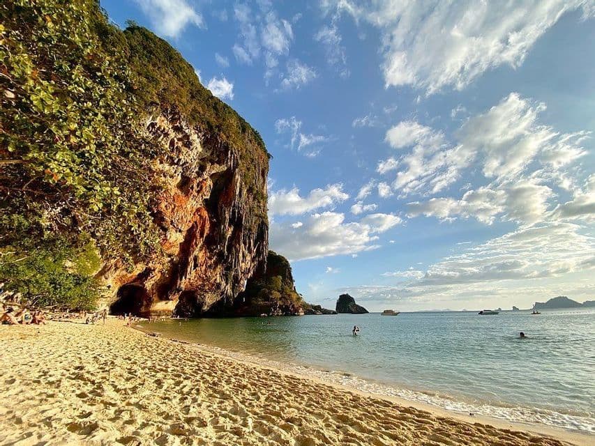 Una grande scogliera rocciosa con una grotta delimita una spiaggia sabbiosa dove le persone nuotano e si rilassano sotto un cielo parzialmente nuvoloso.