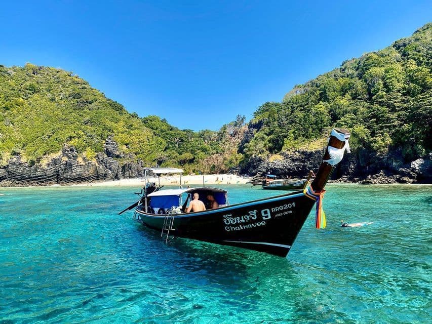 Una tradizionale barca long-tail con a bordo viaggiatori di un gruppo WeRoad galleggia su acque calme e turchesi vicino a un'isola tropicale con spiaggia di sabbia.