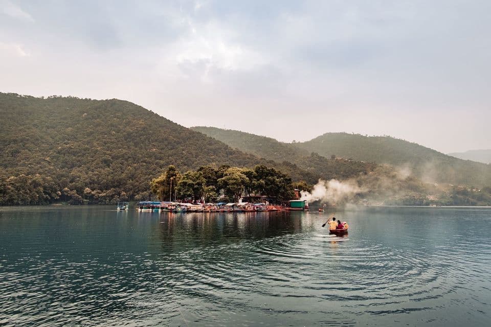 Dos personas reman en un pequeño bote en un lago hacia una pequeña isla, con grandes colinas arboladas al fondo bajo un cielo nublado.