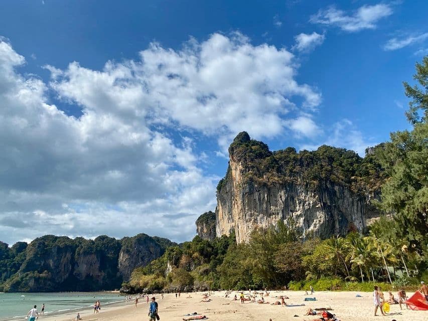 Persone si rilassano su una spiaggia sabbiosa ai piedi di grandi scogliere calcaree sotto un cielo azzurro con nuvole bianche.