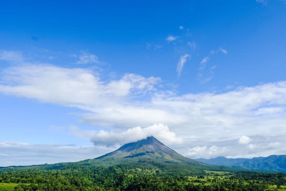 Un grande vulcano con la cima avvolta dalle nuvole, che si erge sopra una fitta foresta verde sotto un cielo azzurro brillante.