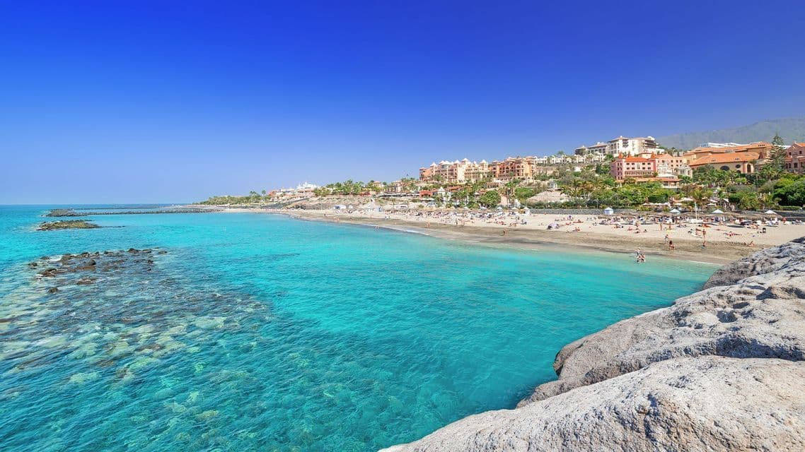 Ein weiter Blick auf einen überfüllten Sandstrand mit klarem türkisfarbenem Wasser, mit einer Ferienortschaft auf einem Hügel unter tiefblauem Himmel.