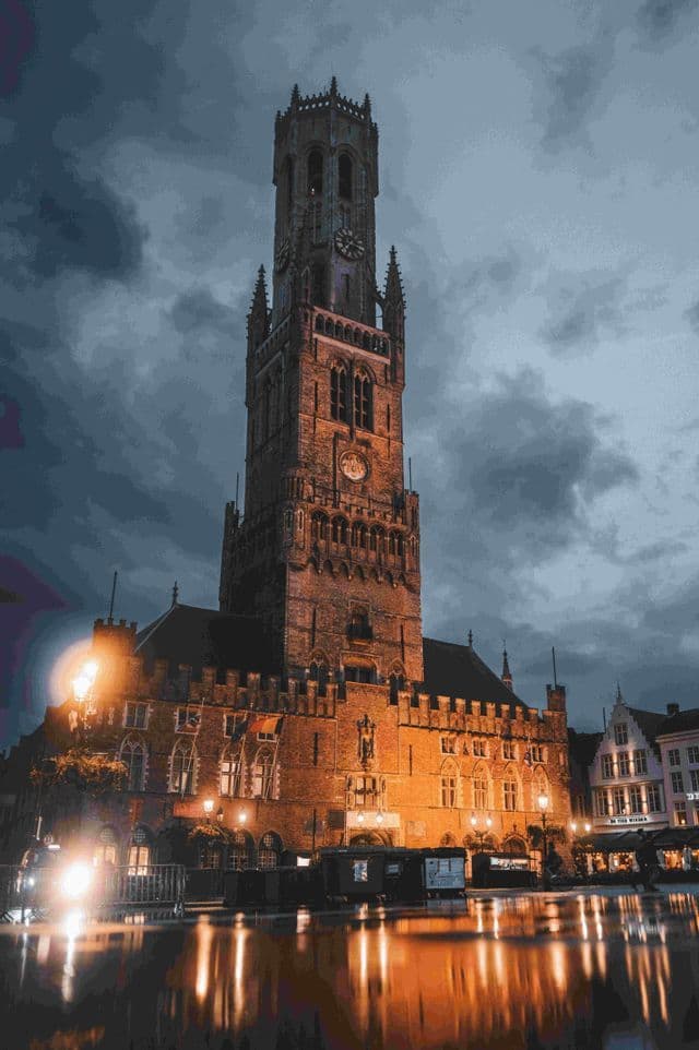 Un'alta, storica torre dell'orologio in mattoni illuminata di notte, le sue luci calde che si riflettono su una piazza cittadina bagnata.