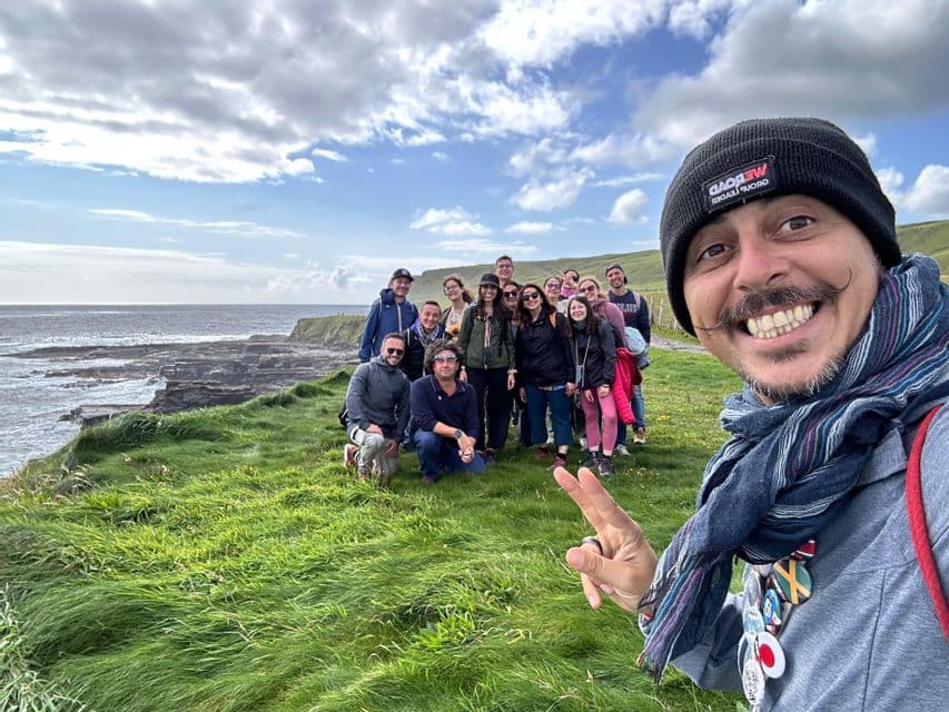 Ein Mann macht ein Selfie mit seiner WeRoad-Reisegruppe auf einer grünen Klippe, im Hintergrund das Meer und ein bewölkter Himmel.