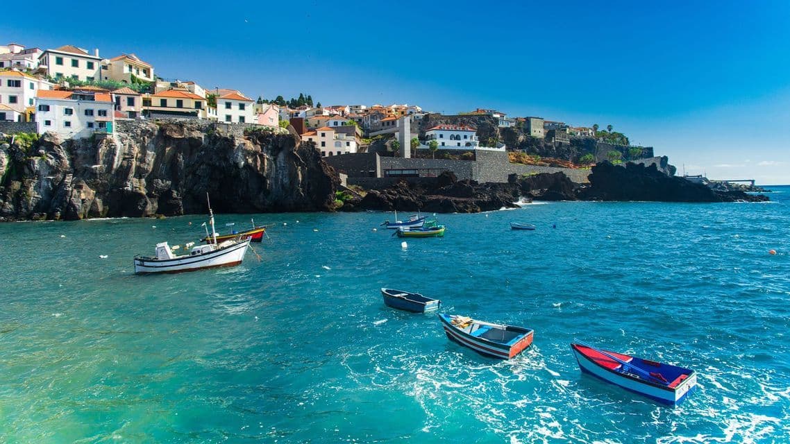 Several small fishing boats float in a turquoise bay in front of a village built on a rocky cliff.