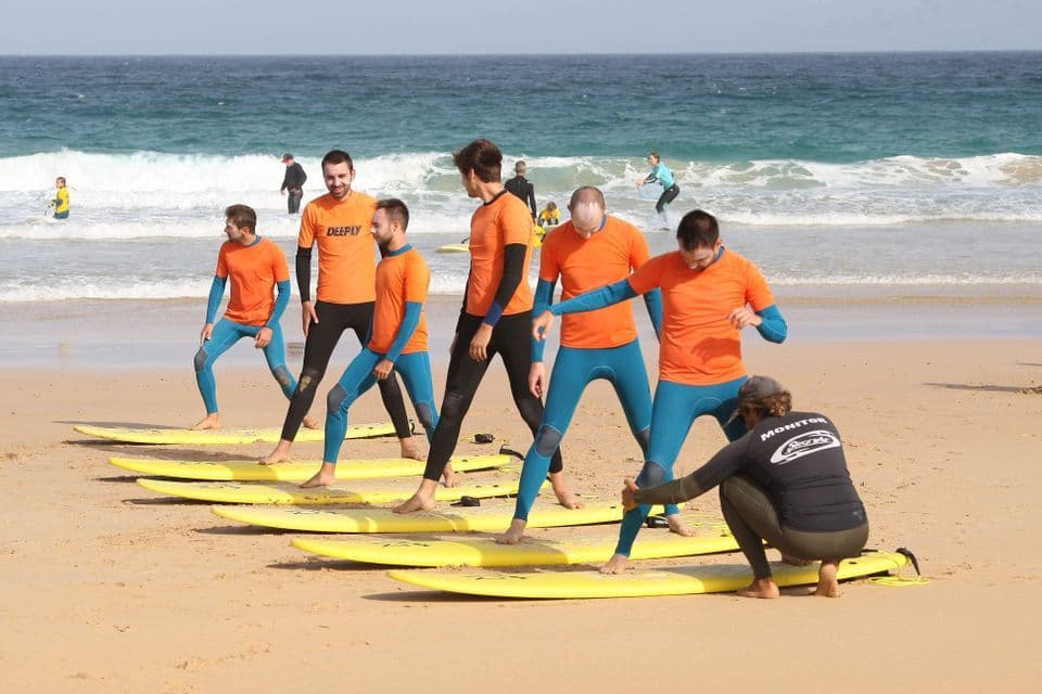 A WeRoad group trip in wetsuits learns to surf from an instructor on a sandy beach with yellow surfboards.