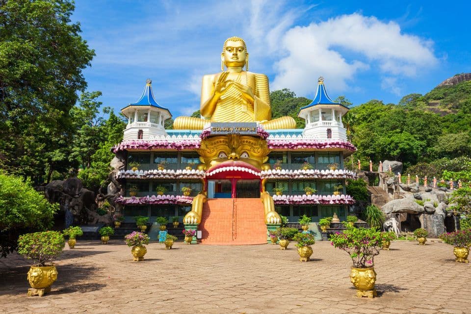 Una enorme estatua dorada de Buda se asienta sobre un templo ornamentado con escaleras que conducen a una entrada con forma de boca de un gran monstruo.