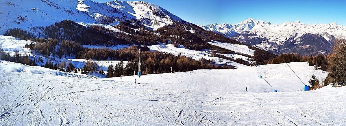 Vista panoramica di un'ampia pista da sci battuta in una giornata di sole, con montagne e foreste innevate sullo sfondo.