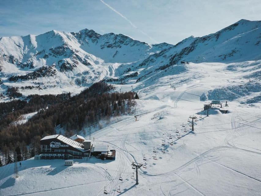 Veduta aerea di una stazione sciistica con rifugio e seggiovie su una montagna innevata sotto un cielo azzurro.