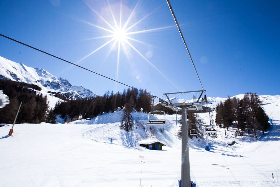 Una seggiovia si muove su un paesaggio montano innevato con pini sotto un sole splendente e un cielo azzurro limpido.