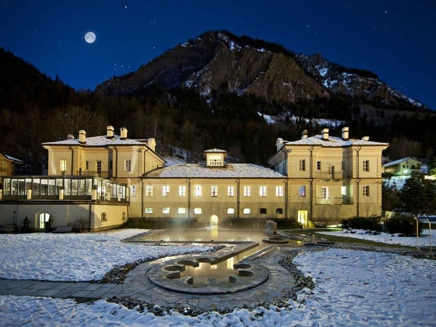 Un edificio illuminato con una piscina termale esterna in un paesaggio innevato, con una grande montagna e una luna piena nel cielo notturno.