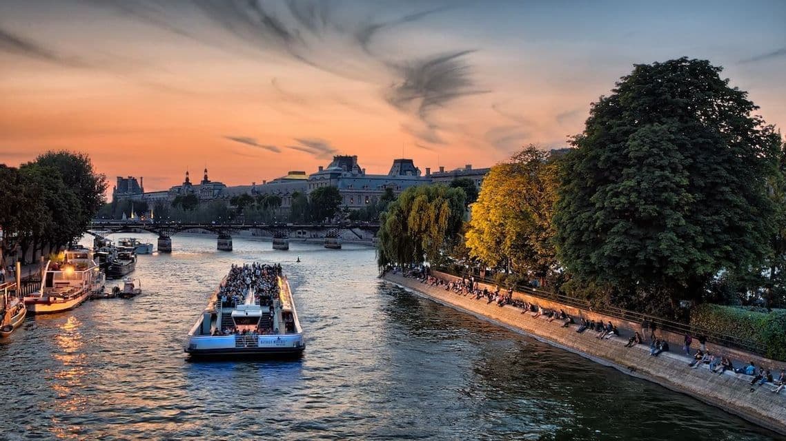 A tour boat full of people sails on a river through a city at sunset, as others relax on the riverbank.