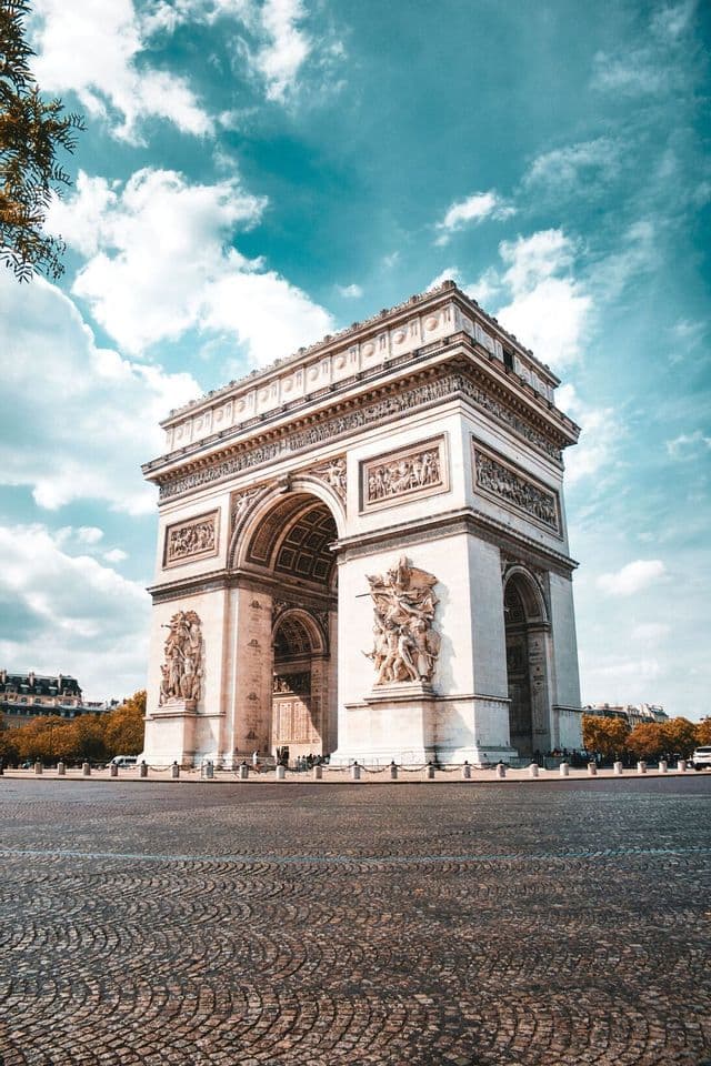 A large triumphal arch with intricate carvings stands on a wide cobblestone plaza under a partly cloudy sky.