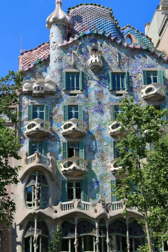 Una facciata di un edificio coperta di mosaici colorati, con balconi a forma di teschio e un tetto a scaglie sotto un cielo blu.