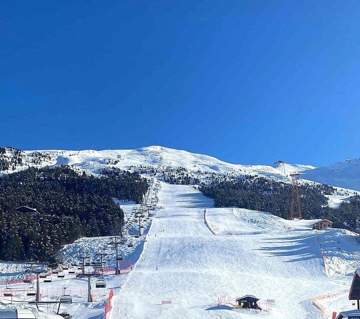 Un'ampia pista da sci con seggiovie in funzione risale una montagna innevata sotto un cielo azzurro chiaro.