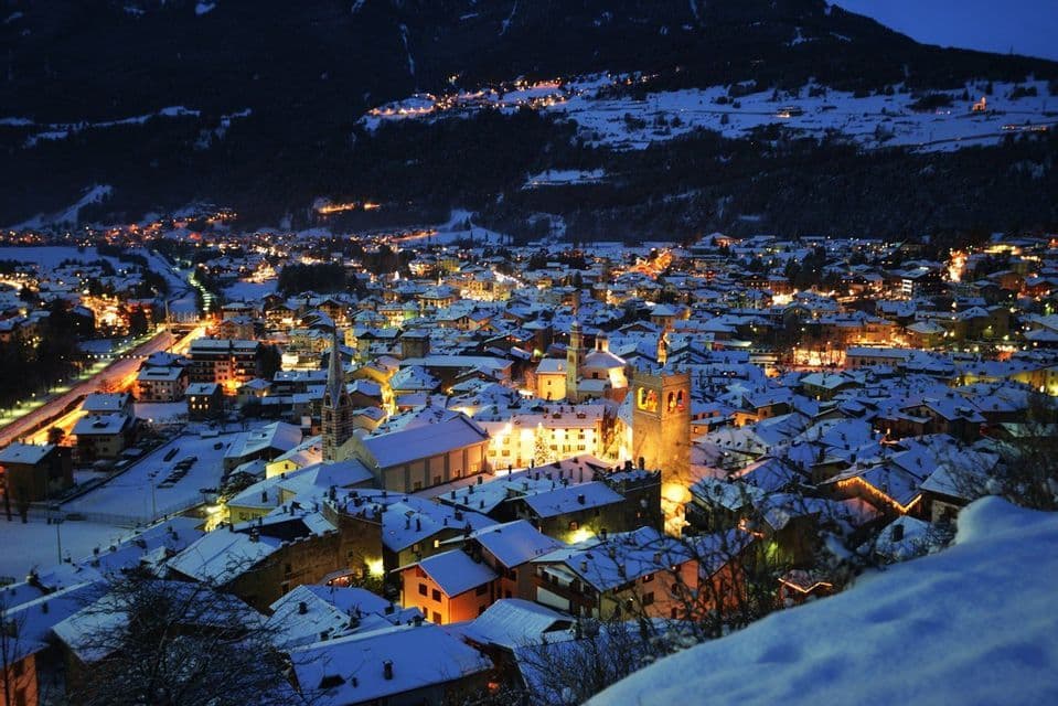 Un villaggio innevato visto dall'alto al crepuscolo, con i suoi edifici che brillano di luci calde contro una montagna scura.