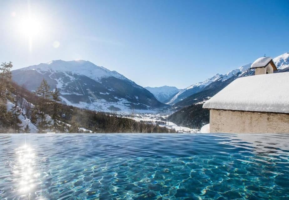 Il bordo di una piscina a sfioro con acqua cristallina si affaccia su una valle montuosa innevata sotto un sole splendente.