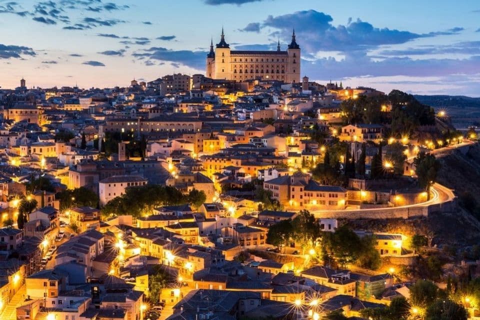 Una città storica su una collina si illumina di luci calde al tramonto, con una grande fortezza che si erge nel punto più alto.