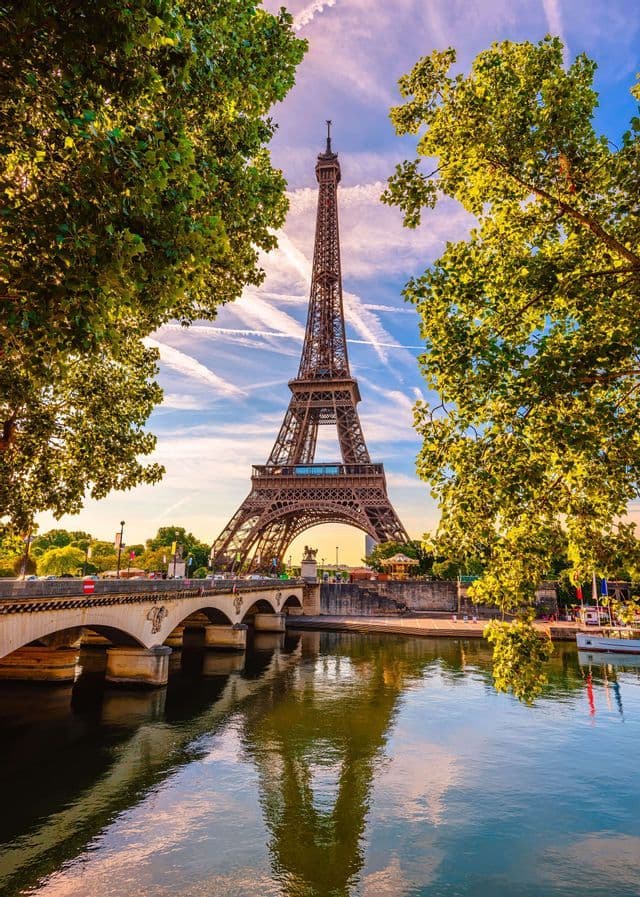 The Eiffel Tower seen from across a river, framed by green trees with its reflection visible in the water near a bridge.
