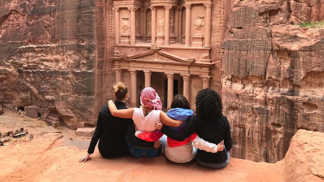 A WeRoad group trip of four women sitting with their arms around each other, looking at an ancient temple carved into a rock cliff.