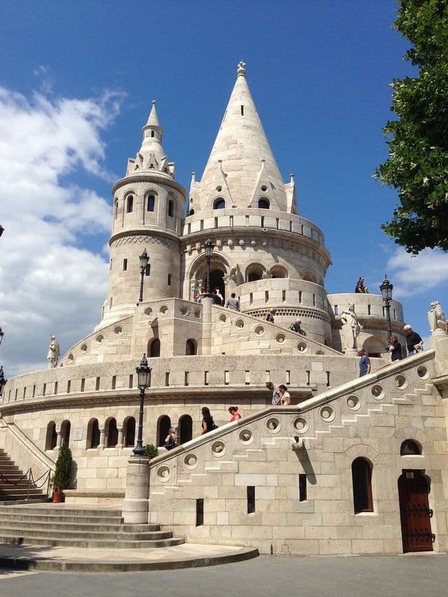 Des touristes marchent sur les escaliers en pierre sinueux d'une grande forteresse blanche aux tours coniques sous un ciel bleu.