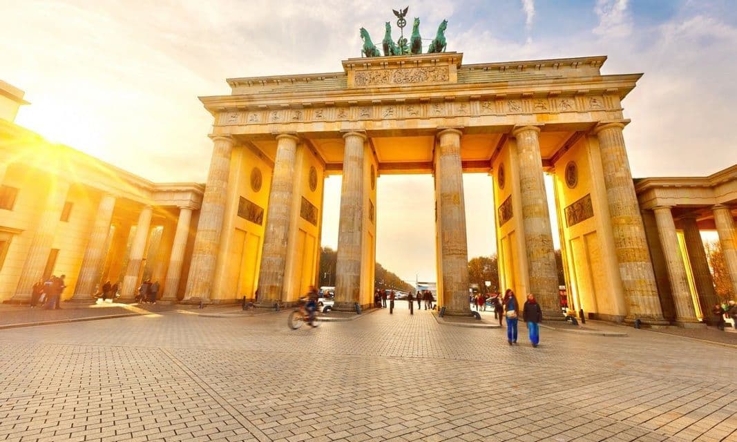 The Brandenburg Gate monument illuminated by a golden sunset, with people walking and cycling on the cobblestone plaza in front.