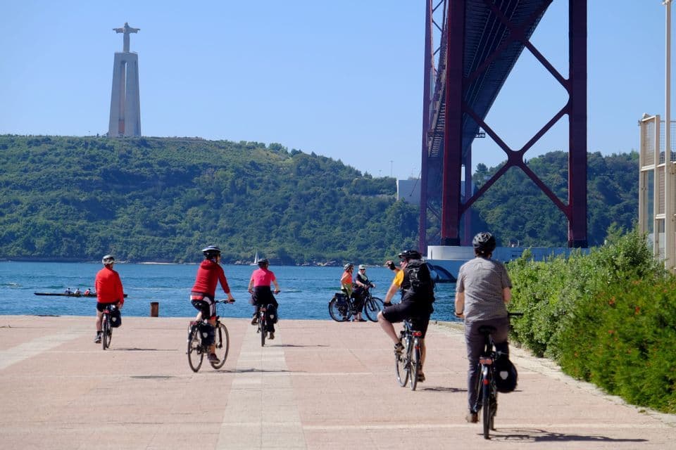 Un viaggio di gruppo WeRoad in bicicletta lungo un percorso lungomare, con un grande ponte rosso e un monumento su una collina distante.