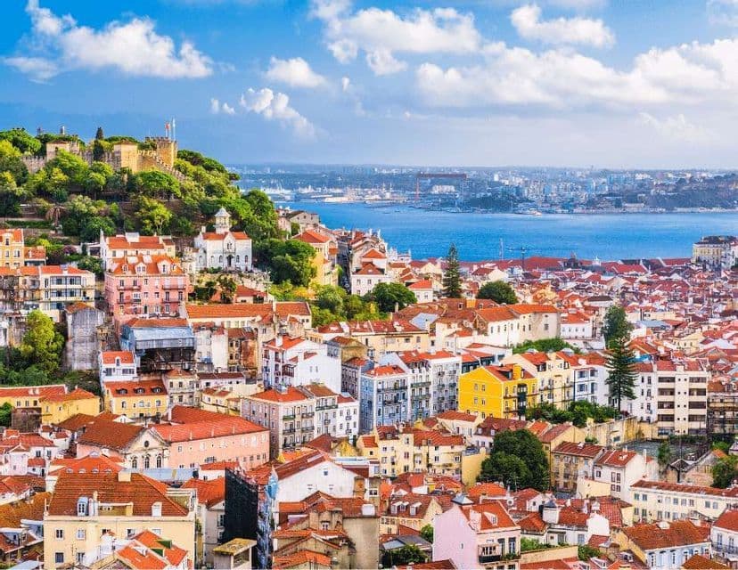 Una vista panoramica di una colorata città costiera con tetti rossi, un castello su una collina e un grande specchio d'acqua sotto un cielo blu.