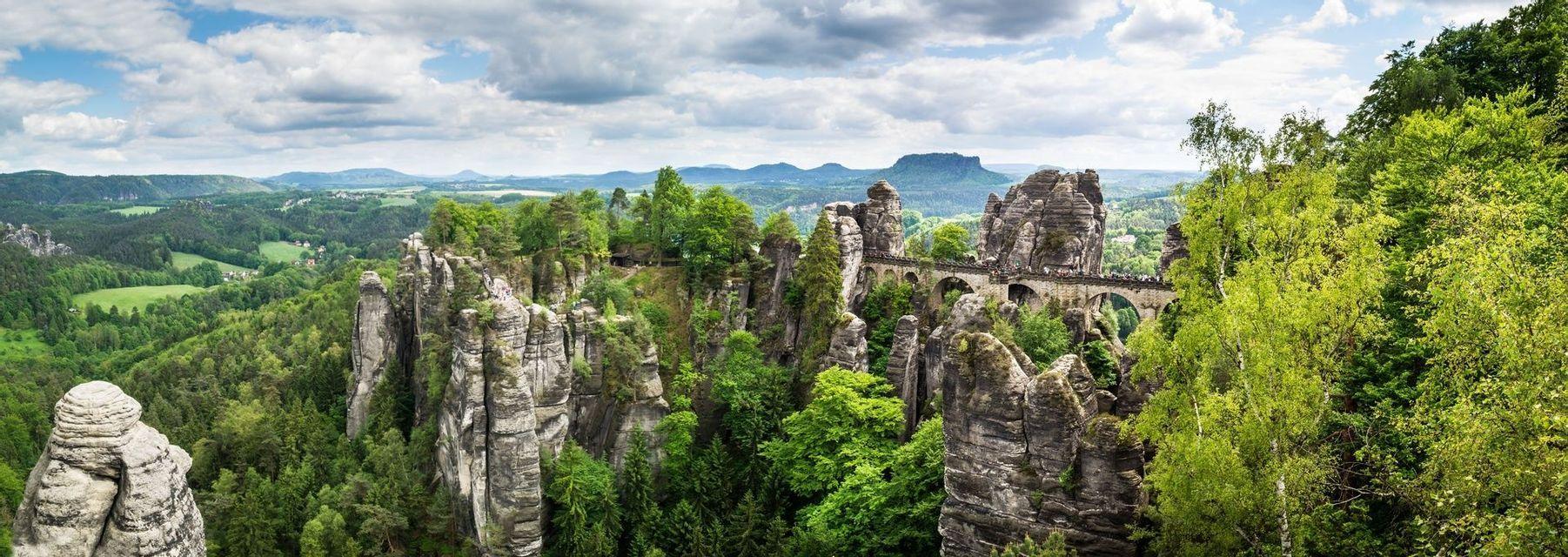 A panoramic view of an arched stone bridge connecting towering rock formations over a vast green forest landscape under a cloudy sky.