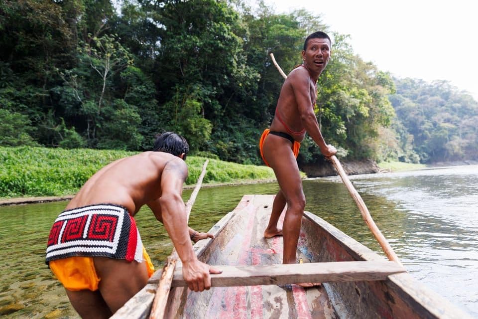 Due uomini in perizomi tradizionali remano su una canoa di legno in un fiume, con una fitta giungla verde sulla riva.