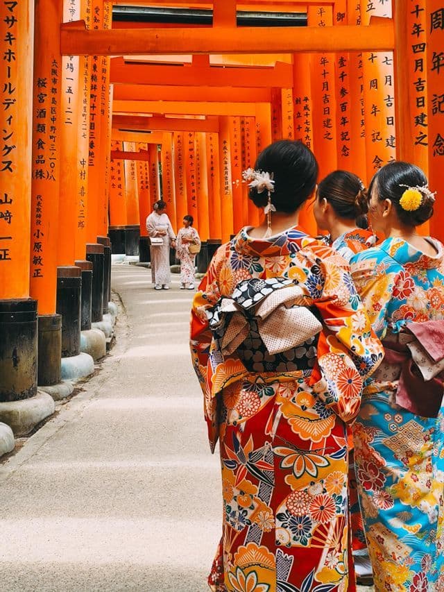 Eine WeRoad-Gruppe von Frauen in farbenfrohen Kimonos spaziert einen Pfad entlang, der von orangefarbenen Torii-Toren gesäumt ist.